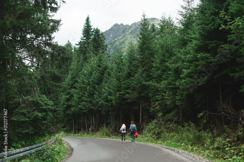 Two female tourists are walking on an asphalt road in the mountains. Travel in the mountains concept, walk on the road. Background. Hiking with girlfriend.