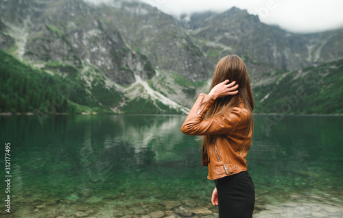 The back of a fashionable girl in a brown leather jacket and skirt stands on a lake in the mountains and poses for the camera. Stylish woman at Morskie Oko Lakes in Tatra Mountains