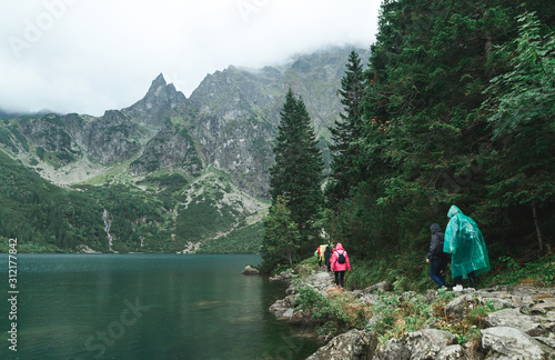 Fototapeta Naklejka Na Ścianę i Meble -  Group of tourists in raincoats walk along a rock path near beautiful Morskie Oko Lake with clear water against a rocky Tatra Mountains background. Tourists and road hiking in the rain.