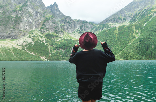 Back of a stylish girl in dark clothing on a background of a mountain lake with blue water and mountains with clouds. Background. Hipster tourist woman on the shores of Lake Morskie Oko