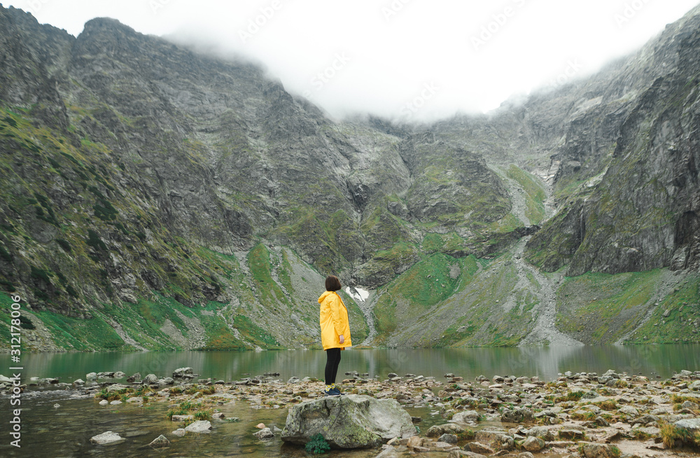 Girl in a yellow jacket stands on a stone against the background of a mountain lake with clear water and looks at the beautiful landscape. Hiker and lake on top of mountain. Background.