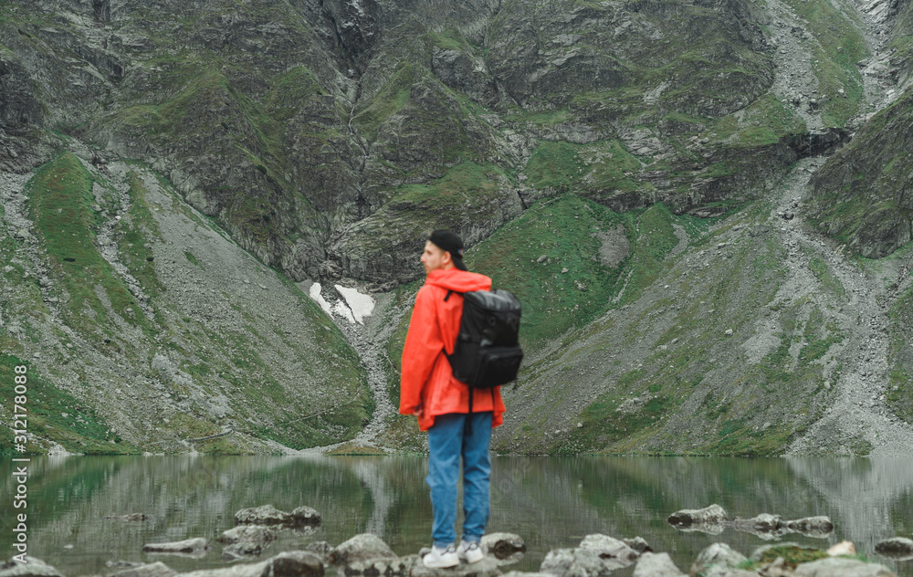 Hiker in a red jacket stands on a rock on the shore of a mountain lake,looks away.Blurred photo of young tourist man with backpack on hike.Abstract creative portrait in defocus on mountains background