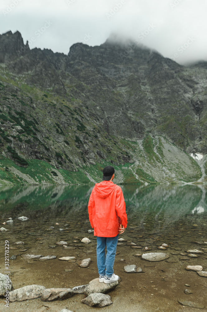 Full length portrait of a young man hiking, standing on top of a mountain near a lake and looking at the beautiful landscape. Vertical photo of tourist in red jacket in mountains near lake. Copy space