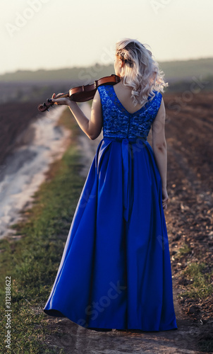 woman in long dress playing violin on background of field path with a club of dust, girl engaged in musical art, performance on nature, concept passion in music