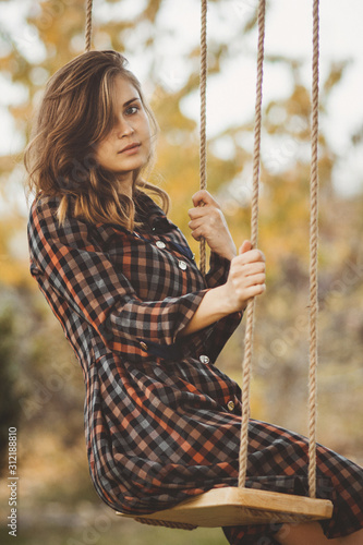 upset girl in a dress sways on a swing in the autumn garden, a young woman in melancholy mood thinkng on nature photo