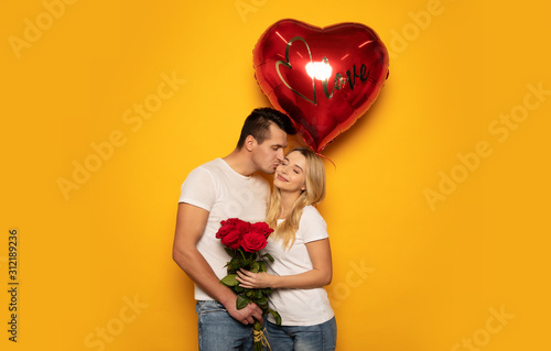 Perfect love. Сlose-up photo of a charming lady and her handsome boyfriend, who is kissing her in the cheek, while giving her a bouquet of roses and a big red heart-shaped balloon.