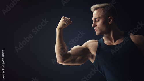 Leinwand Poster Young confindent muscular bodybuilder guy demonstrates biceps on black back
