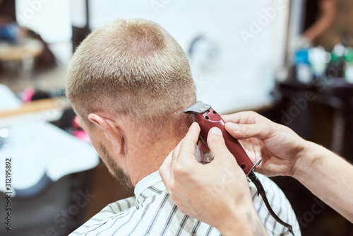 Dark blonde man at barber shop is having his nape shaved with a trimmer, hairdresser is holding a red tool, stripe cape on
