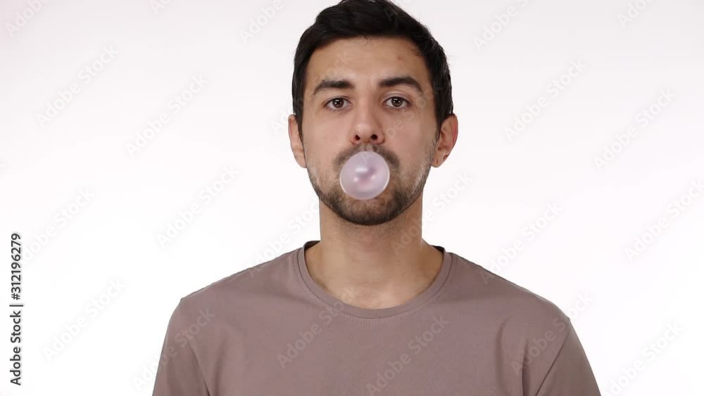 Front view of european dark haired young bristled man blowing chewing gum bubble with eyes wide open isolated over white background. Chewing gum bursts. Slow motion