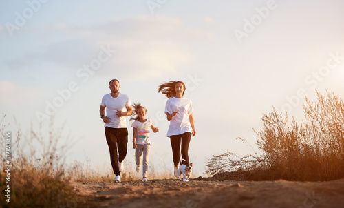 Front view of young and beautiful family of three jogging with their dog outside the city on the village road on the setting sun