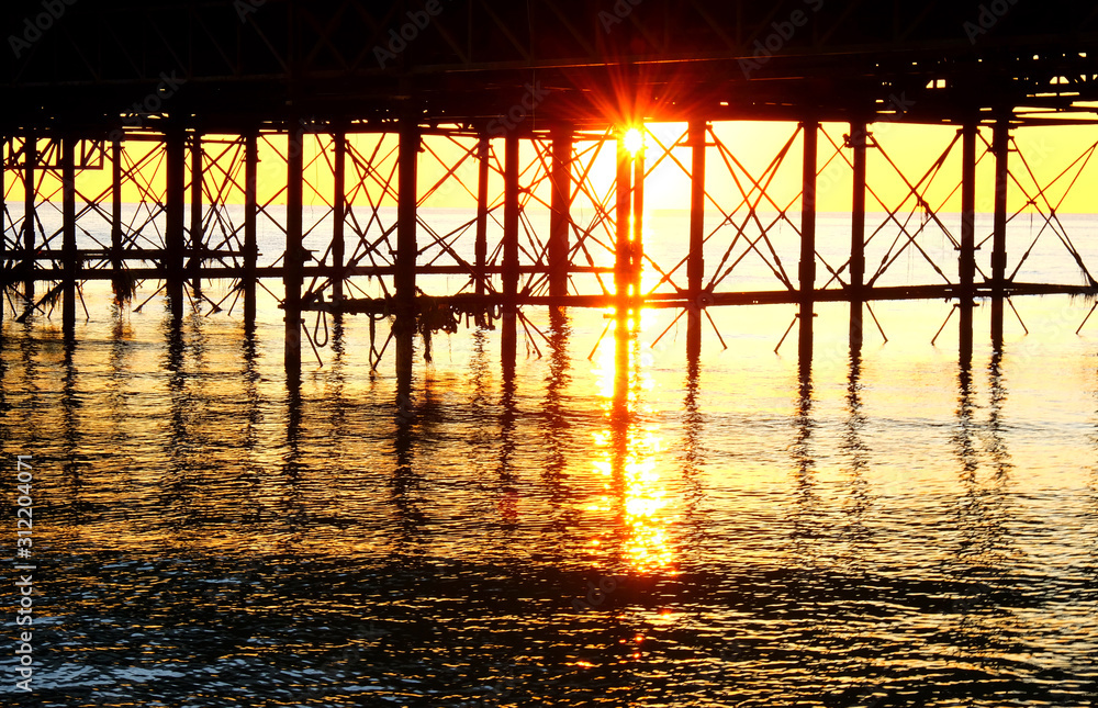 The metal support strutts of Brighton Pier glowing yellow with the sun setting underneath it