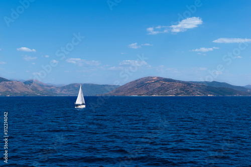 The sea is a classic blue color and a white sailboat on it. Mountains of Greece and the sky in the background. Amazing sea landscape. Beautiful view from the port of Lefkimmi. Corfu island, Greece.