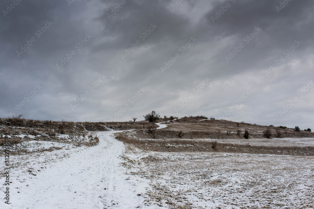 Curving dirt road leading to the top of the hill at wintertime, dramatic storm clouds in background.