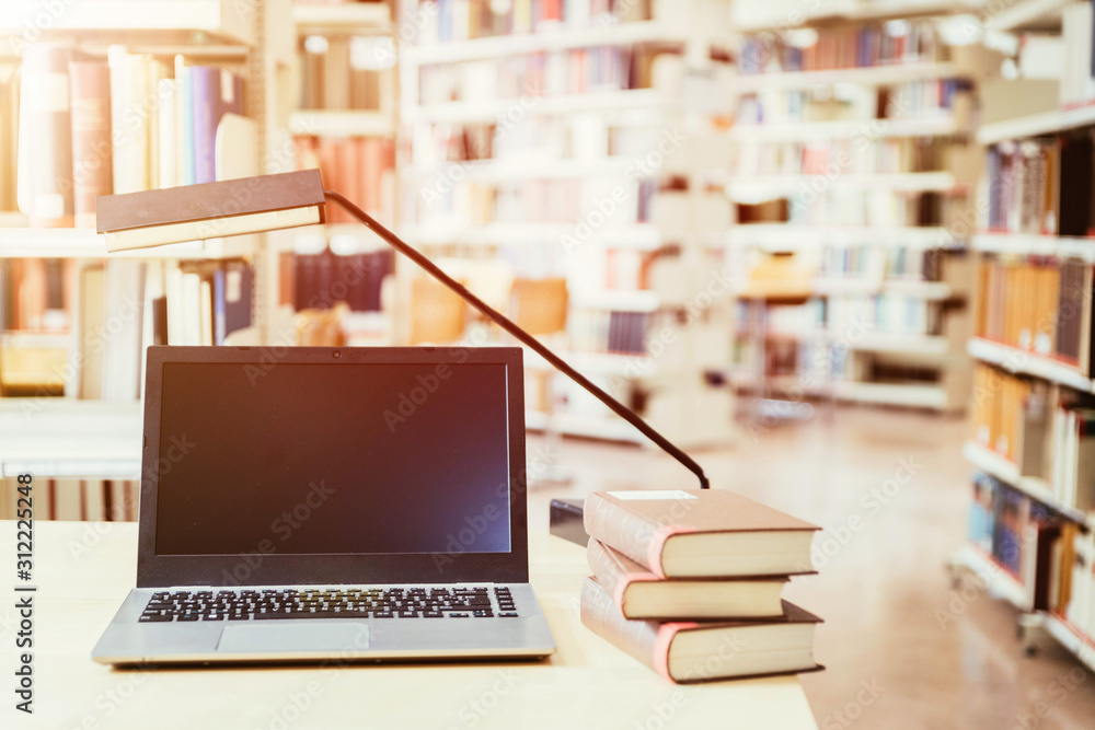 Studying in the university: Laptop on a wooden desk, pile of books and books in the blurry background Studying in the university: Laptop on a wooden desk, pile of books and books in the blurry background