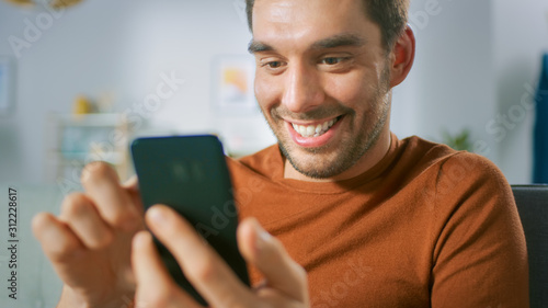 Handsome Excited Happy Man at Home Uses Smartphone. Man Relaxing with Mobile Phone in His Cozy Living Room.