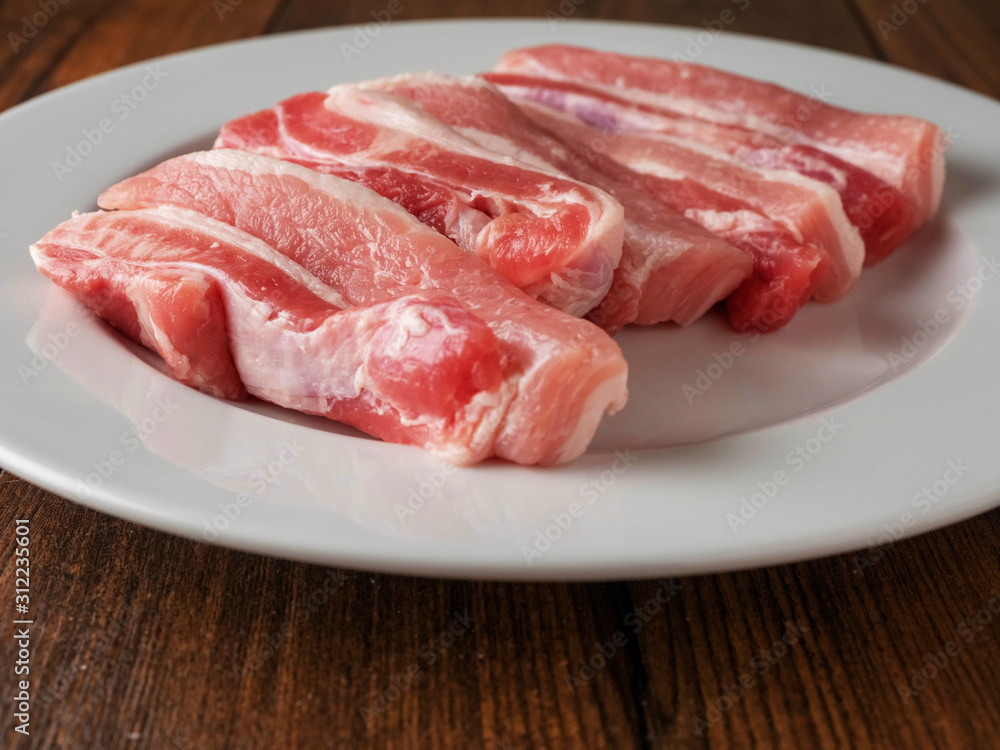 Uncooked raw pork belly slices on a white plate and wooden table.