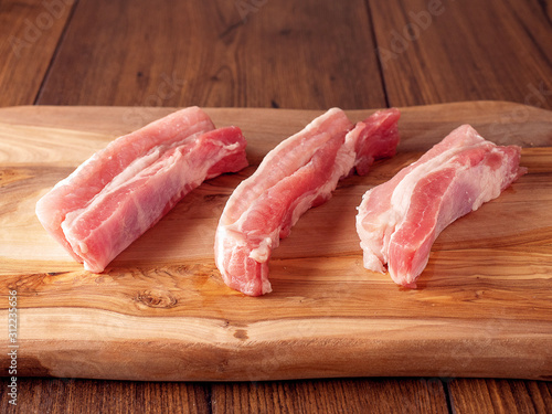 Three raw pork belly slices on a wooden cutting board, Dark wood table surface.