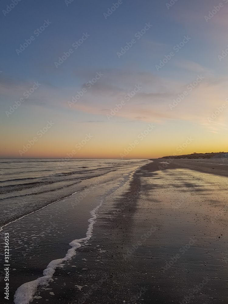 Fototapeta premium Coastline at sand beach with orange colors from the sun in the horizon. View lengthwise of the shore. Sæby, Denmark.