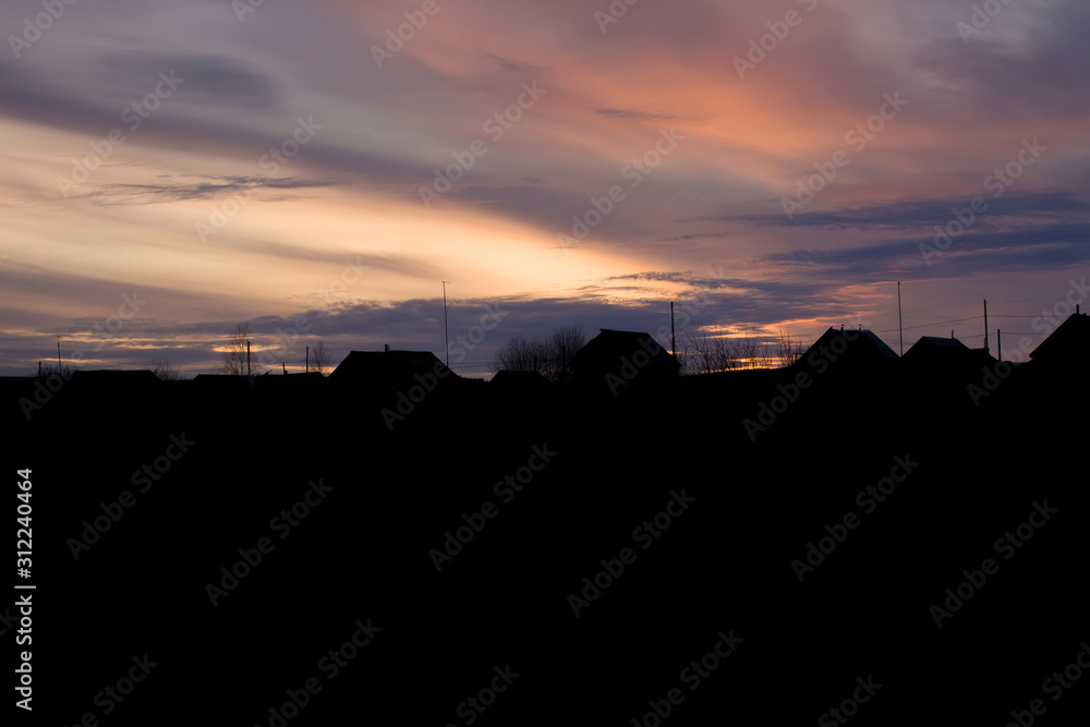 Obraz premium The silhouette of houses and roofs against the sunset. Rural houses and trees on the background of a colorful gorgeous sunset.