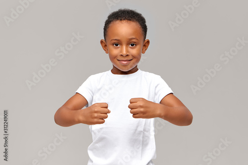 Studio shot of funny ten year old African boy in white t-shirt keeping clenched fists in front of him demonstrating strength or holding invisible objects, looking at camera with playful smile