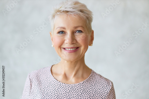 Close up image of good looking beautiful mature blonde female with blue eyes, elegant make upand pixie hairstyle smiling at camera, having confident happy facial expression, being in good mood