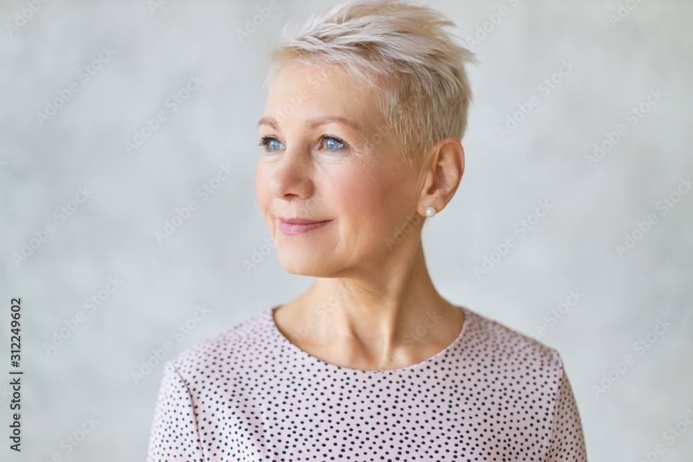 Close up studio image of beautiful attractive middle aged European lady with stylish haircut and neat make up looking away with confident smile posing isolated against marbled wall background - obrazy, fototapety, plakaty 