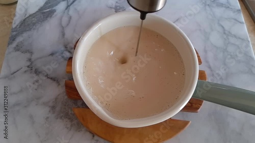 Frothing plant milk for morning latte coffee in an enamel saucepan with electric frother. Top view. Slow motion