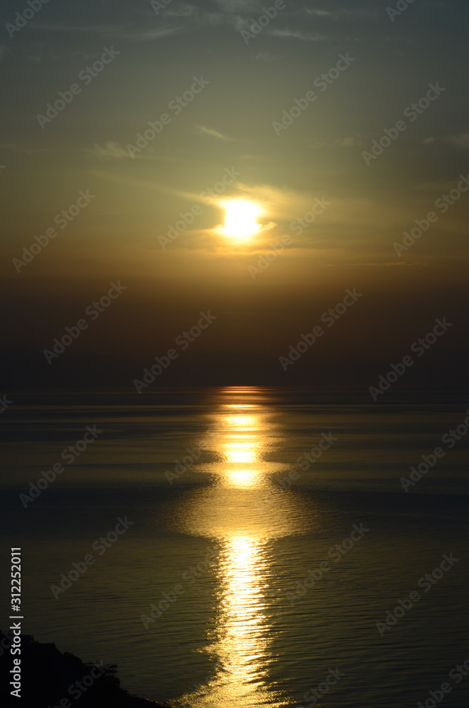 Highly bright sun over the calm sea, adorned with a long&wide solar track on slightly rippled water. Contrast seascape: a glowing sun behind the rare light clouds, above the deeply shadowed horizon