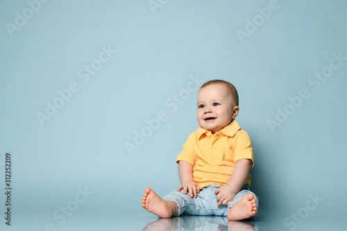 Smiling baby boy in jeans and yellow shirt is sitting on the floor, listening something interesting at free copy space