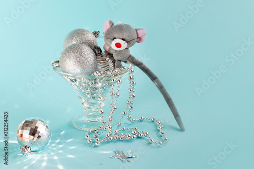 The wide glass is filled with silver Christmas balls and beads, gray toy mouse sits on top - symbol of 2020. Shiny ball with many faces reflects light on a blue background. Copy space photo