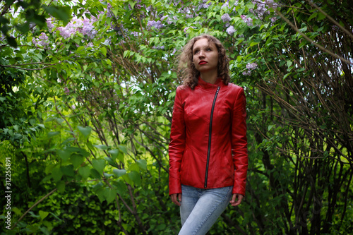 Frontal portrait of a beautiful smiling brunette girl, wear in red leather jacket, looking at you against autumn green, yellow, sunset background.