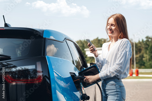Transportation. Young woman on electric car having stop at charging station standing plugging cable browsing smartphone joyful while charging