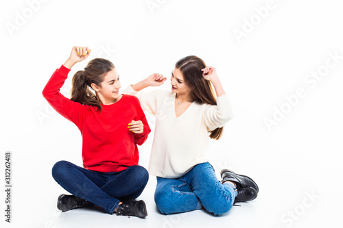 Two girls friends sitting on the floor with win gesture isolated on white background