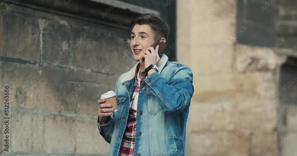 Caucasian young stylisg guy student walking the street and speaking on the mobile phone, sipping coffee to-go. Outside. Caucasian young stylisg guy student walking the street and speaking on the mobile phone, sipping coffee to-go. Outside.