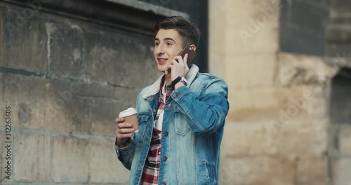 Caucasian young stylisg guy student walking the street and speaking on the mobile phone, sipping coffee to-go. Outside. photo