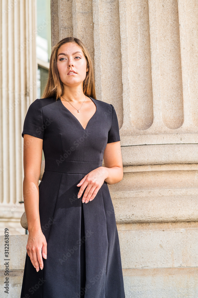Portrait of Young East European Businesswoman in New York. Young woman with long brown hair, wearing black short sleeve, v neck dress, standing by column outside office building, looking forward..