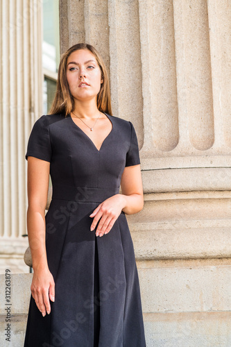 Obraz na płótnie Portrait of Young East European Businesswoman in New York. Young woman with long brown hair, wearing black short sleeve, v neck dress, standing by column outside office building, looking forward.. w salonie