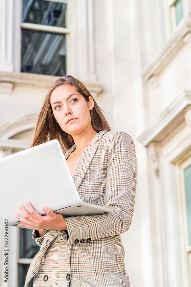 Way to Success. Young East European woman with long brown hair, wearing patterned blazer, holding laptop computer, standing outside vintage office building in New York City, looking away, thinking. Way to Success. Young East European woman with long brown hair, wearing patterned blazer, holding laptop computer, standing outside vintage office building in New York City, looking away, thinking.