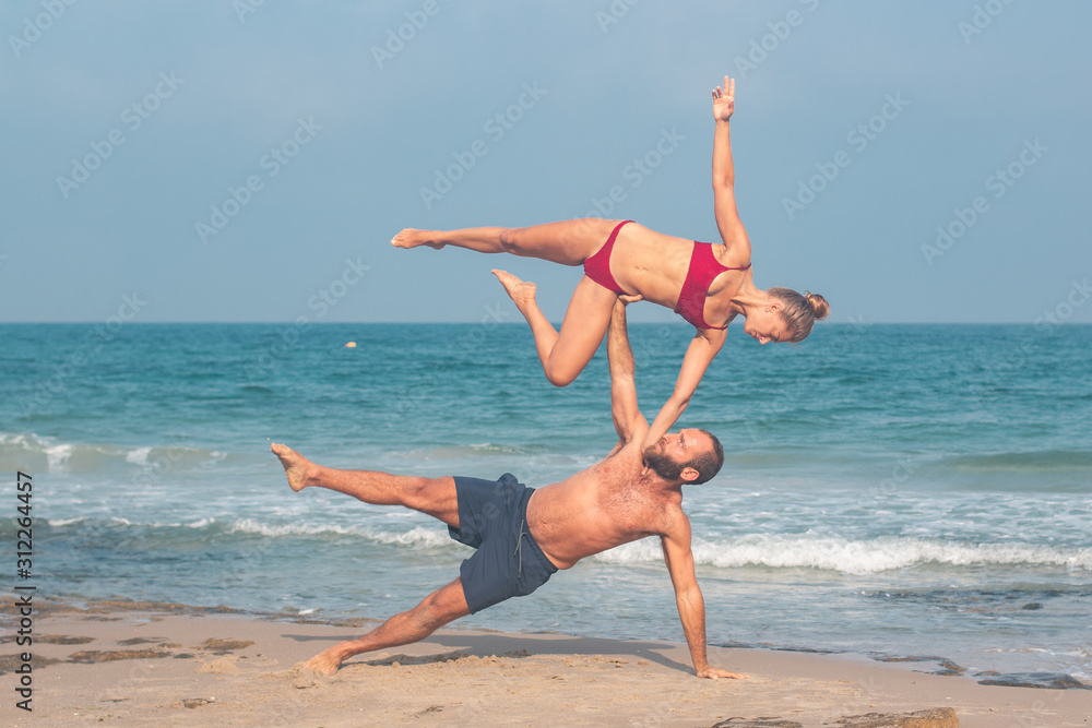 Muscular man and fit young woman training together in Acro Yoga. Morning sun lighting the couple and the mediterranean beach background.	