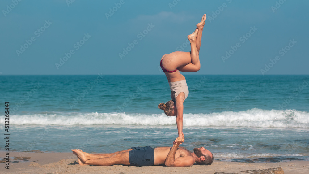 Muscular man and fit young woman training together in Acro Yoga. Morning sun lighting the couple and the mediterranean beach background. Muscular man and fit young woman training together in Acro Yoga. Morning sun lighting the couple and the mediterranean beach background.