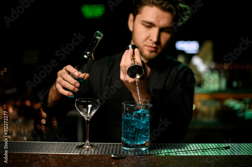 Professional male bartender pouring a blue alcoholic liquor from the jigger to a measuring cup