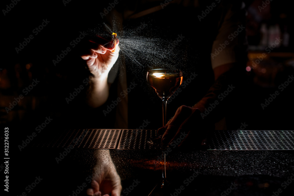 Professional male bartender spraying on the brown alcoholic drink with a orange zest juice in the dark