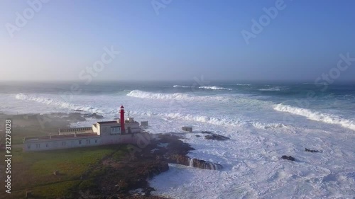 Aerial, descending, drone shot of the Cabo Raso lighthouse, in front of big, stormy waves crushing the Portuquese coast, after the Fabian storm, on a misty and sunny morning, in Portugal photo
