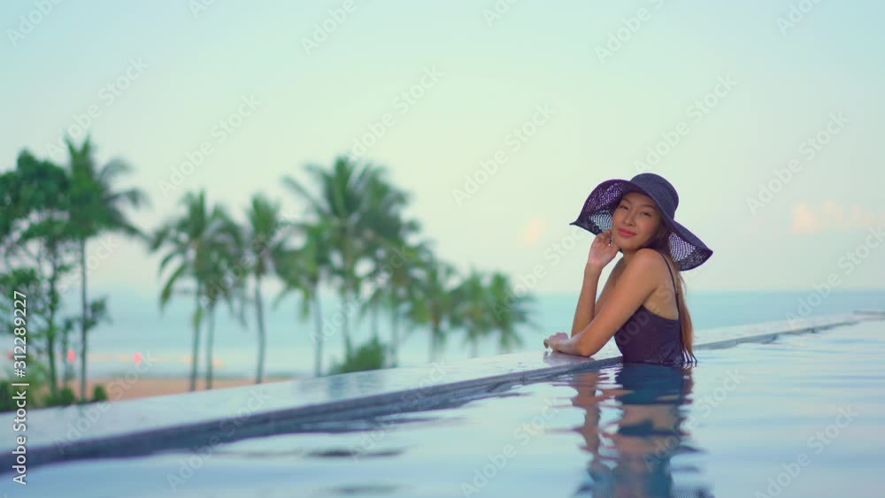 locked medium-wide portrait shot of young adult Asian woman in swimsuit with hat sitting at the edge of infinity pool with defocussed tropical beach and palm trees background copy space locked medium-wide portrait shot of young adult Asian woman in swimsuit with hat sitting at the edge of infinity pool with defocussed tropical beach and palm trees background copy space
