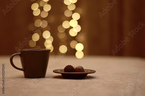  A brown cup and saucer with bright lights, above the cup, and a plate of chocolate candies, sprinkled with cocoa.