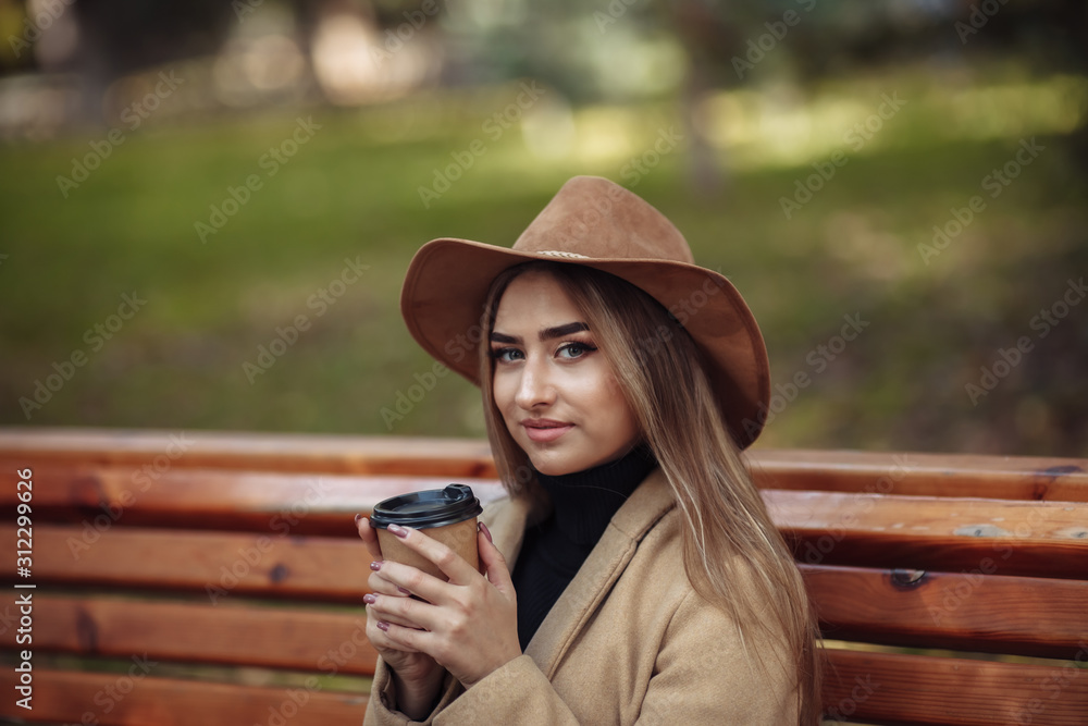 Young attractive woman in autumn clothes sits on bench and drinks coffee in city park. Woman dressed in stylish coat and felt hat. Autumn time
