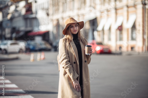 Young stylish woman dressed in a coat and felt hat walks around the city and drinks coffee on the go. Autumn time