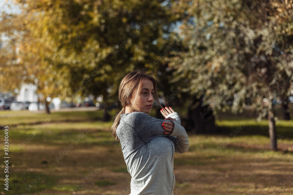 Young sport woman in a sports suit is warming up in the autumn park. Stretching hands before training. Fitness training outdoors