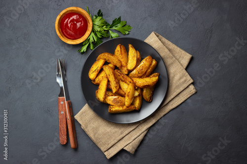 Baked potato with spices, herbs and ketchup in a black plate. Dark background. Top view with copy space.