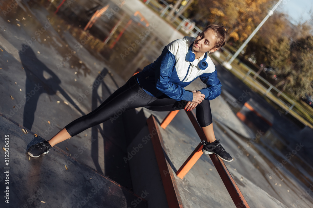 Obraz premium Fit slim woman in sportswear and headphones practicing leg stretching before training in a sports park